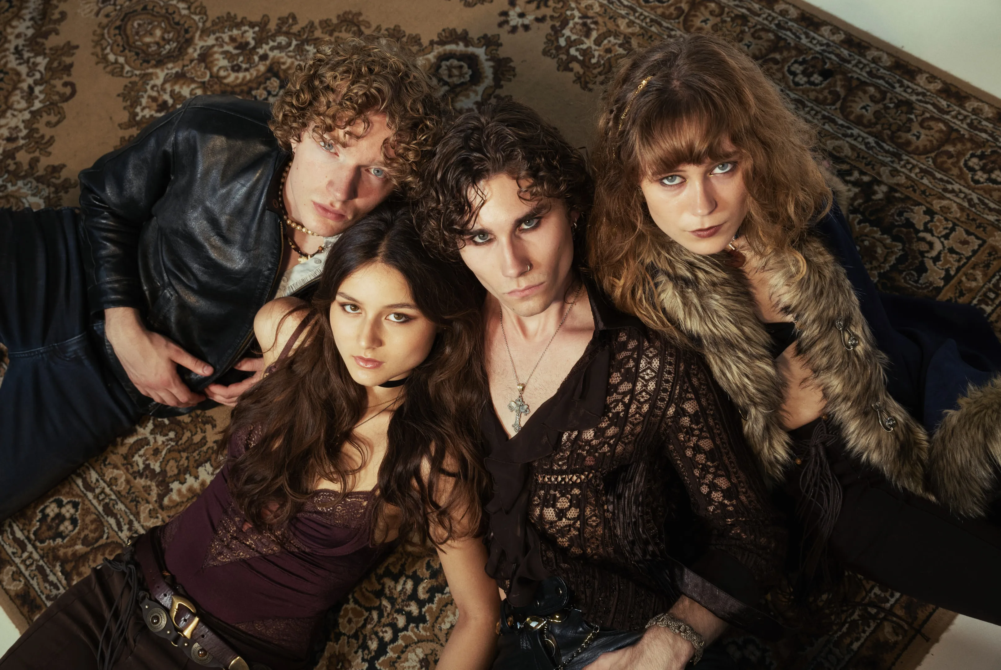 The four members of Corduroy County photographed from above, lying close together on a patterned brown carpet in leather, lace and fur.